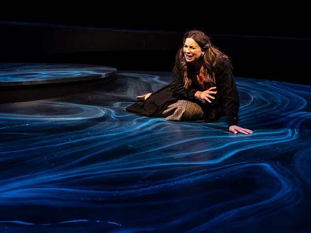A woman is sitting on a swirling blue stage set, upset or crying.